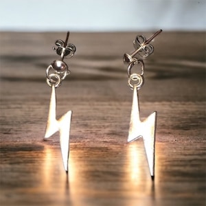 May include: A pair of silver stud earrings with a lightning bolt charm hanging from each. The earrings are on a wooden surface.