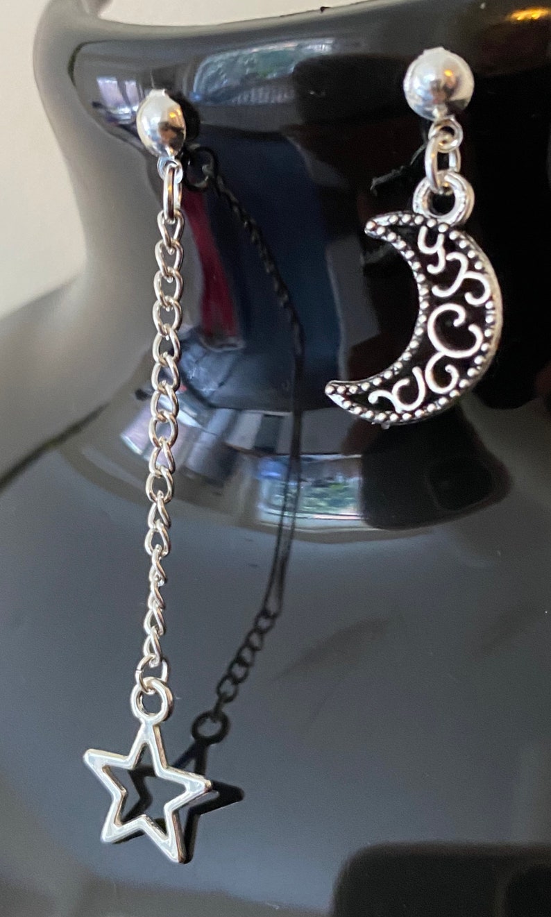 May include: A pair of silver earrings, one with a crescent moon charm and the other with a star charm hanging from a silver chain.