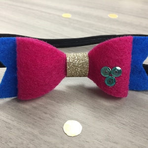 May include: A pink and blue felt bow with a gold glitter centre and teal sequins. The bow is attached to a black headband.
