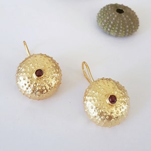 Pode incluir: Um par de brincos de ouro em forma de ouriço-do-mar com uma pequena gema vermelha no centro de cada brinco.
