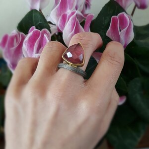 Op de afbeelding: Handgemaakte ring met een grote, facetgeslepen, roodroze edelsteen gezet in een goudkleurige bezel. De ringband is van een getextureerd, zilverkleurig metaal. De achtergrond omvat roze en witte bloemen en groene bladeren.