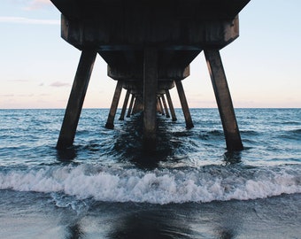 May include: A low-angle shot of a pier extending over the ocean. The concrete structure is supported by large, angled pillars. The water is a deep blue, with waves breaking on the shore. The sky is a pale blue.