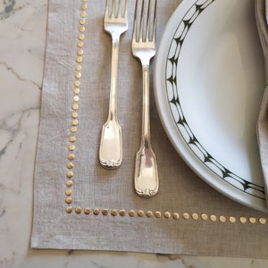 Könnte beinhalten: Ein beigefarbenes Leinen-Platzset mit einem goldenen Punktmuster und zwei silbernen Gabeln darauf. Ein weißer Teller mit einem grün-weißen geometrischen Muster liegt auf dem Platzset.