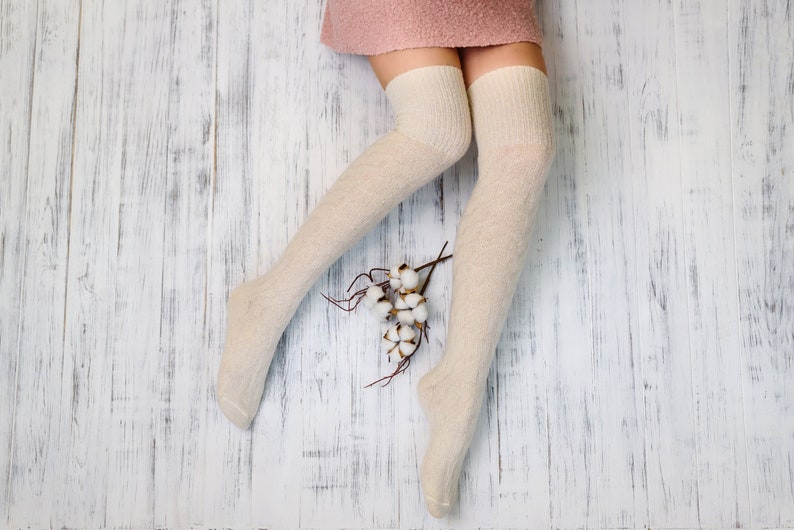 K&ouml;nnte beinhalten: Ein Paar wei&szlig;e Oberschenkelsocken mit einem rosa Rock im Hintergrund. Die Socken sind aus weichem, gestricktem Stoff und haben einen dezenten Glanz. Die Socken liegen auf einem wei&szlig;en Holzboden mit einem kleinen Strau&szlig; wei&szlig;er Baumwollbl&uuml;ten.