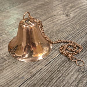 May include: A copper bell with a chain attached. The bell is shiny and has a rounded shape. The chain is made of small, linked copper pieces.