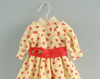 May include: A yellow dress with a red heart pattern and a red ribbon around the waist. The dress has long sleeves and a white lace collar. A small red rose with green leaves is attached to the ribbon.