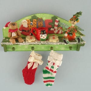 Peut inclure: Une étagère en bois verte sur le thème de Noël. L'étagère a une pancarte qui dit "Jingle" et présente un bonhomme de neige, un Père Noël, un ours et une chaussette. Deux chaussettes rayées rouge et blanc sont suspendues à l'étagère.