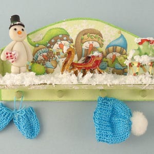 Peut inclure: Une étagère en bois avec une scène d'hiver peinte dessus. Il y a trois bonhommes de neige et un traîneau avec un motif rouge et or. Deux bonnets bleus en tricot sont accrochés à des crochets sur l'étagère.