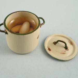 May include: A miniature cream-colored enamel pot with a green rim and two side handles, containing a clear liquid and two white oval-shaped beans. A matching circular lid with a green handle and brown distressed markings sits beside the pot.