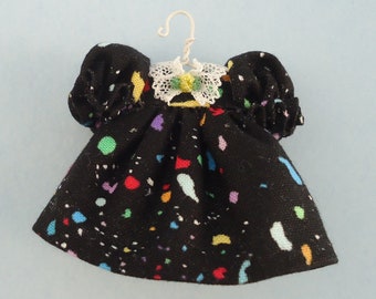 May include: A miniature black dress with colorful confetti-like patterns. The dress features puffy sleeves, a white lace collar, and a small yellow flower accent. It hangs on a white wire hanger.
