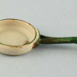 Peut inclure: Une petite poêle jouet de style vintage. L'intérieur est crème, avec un bord et une poignée verts. La poignée se termine par une boucle. La poêle présente des signes d'usure, ce qui ajoute à son charme ancien.