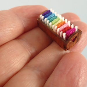 May include: A miniature wooden thread spool holder filled with colorful spools of thread. The spools are arranged in a row, showcasing a rainbow of colors including purple, blue, green, yellow, orange, pink, and red. The holder has a heart-shaped cutout on the side.