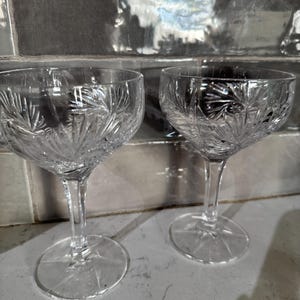 Puede incluir: Dos copas de champán de cristal transparente. Cada copa tiene una base redonda, un tallo delgado y un cuenco ancho con un patrón grabado detallado. Las copas están colocadas sobre una superficie clara contra un fondo reflectante.
