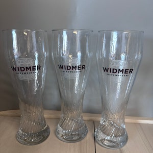 Puede incluir: Tres jarras de cerveza de vidrio transparente con un diseño texturizado. Cada jarra tiene el logotipo de Widmer Brothers Brewing con el texto "Widmer" en negro y "Hefeweizen" en rosa.