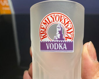 May include: A frosted glass shot glass with a red and purple logo that says "Kreml'ovskaya Vodka".
