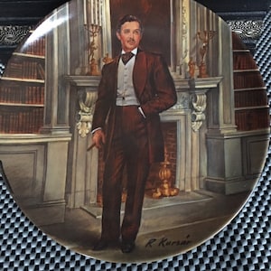May include: A decorative ceramic plate featuring a painting of a man in a formal suit and bow tie, standing in a library setting. The plate is signed "R. Kursar" in the lower right corner.