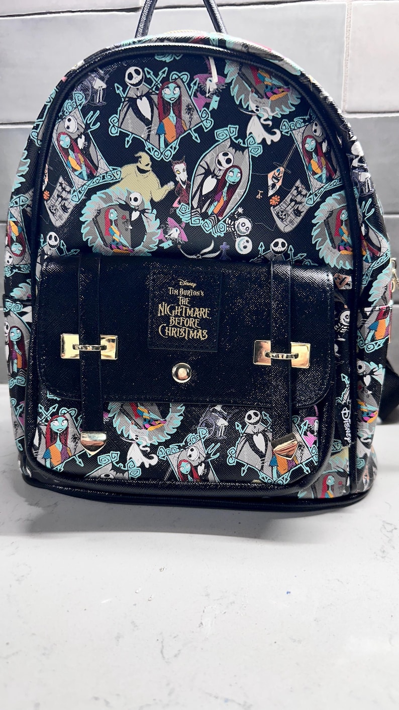 May include: Black backpack featuring a repeating pattern of characters from "The Nightmare Before Christmas." The front flap has a black buckle closure and the text "Tim Burton's The Nightmare Before Christmas."