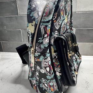 May include: Black backpack featuring characters from The Nightmare Before Christmas. The backpack has a main compartment with a zipper, a smaller front pocket, and a top handle. The design includes Jack Skellington, Sally, and other characters in a repeating pattern.