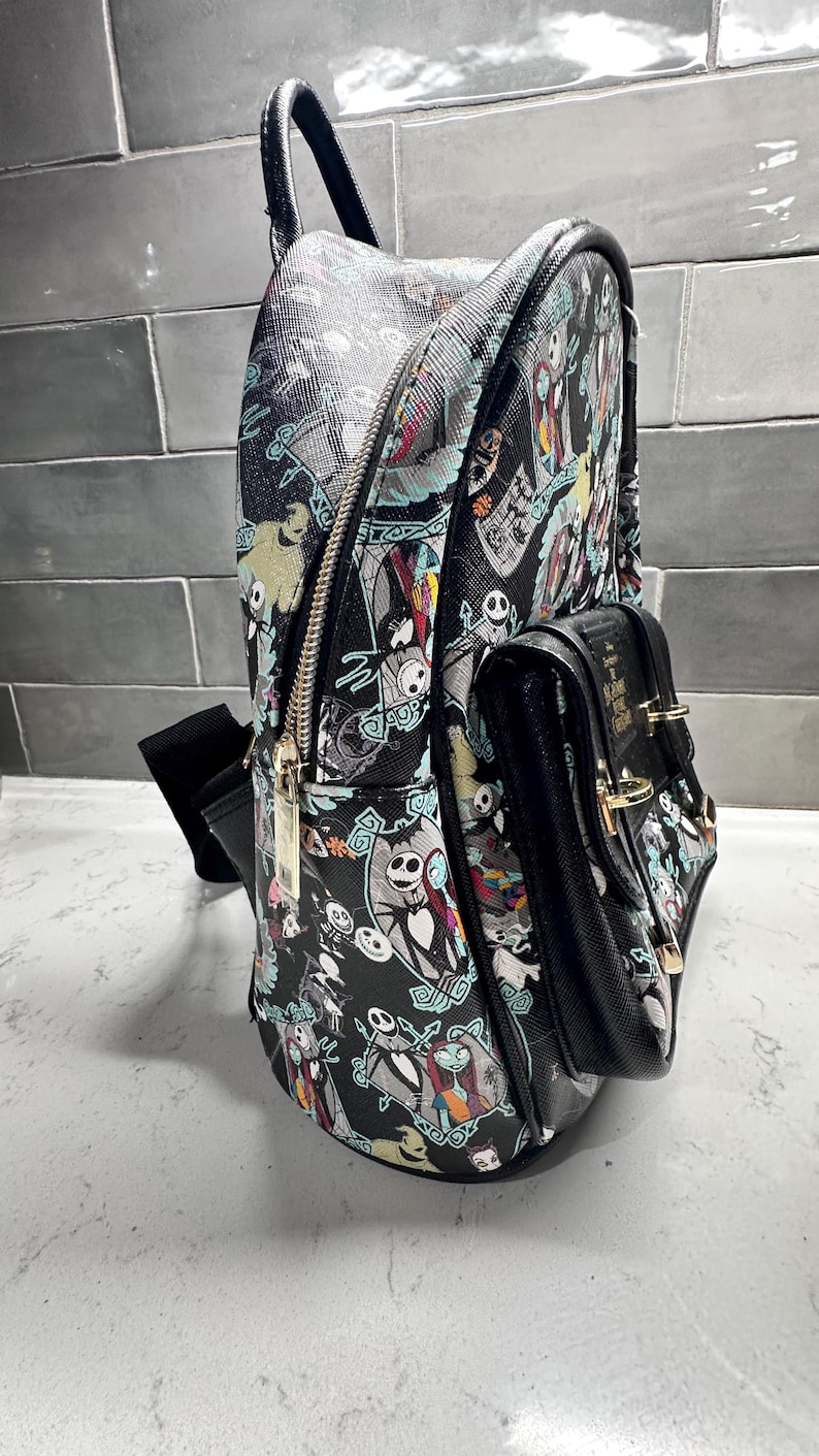 May include: Black backpack featuring characters from The Nightmare Before Christmas. The backpack has a main compartment with a zipper, a smaller front pocket, and a top handle. The design includes Jack Skellington, Sally, and other characters in a repeating pattern.