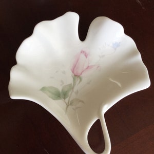 May include: A white ceramic dish shaped like a leaf with a pink rose and green leaves painted on it.