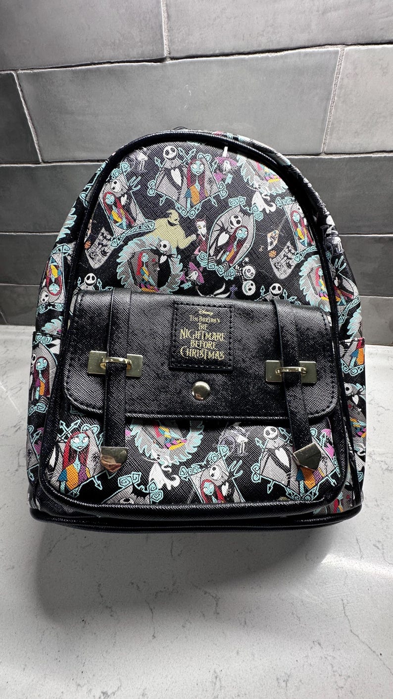 May include: Black backpack featuring characters from "The Nightmare Before Christmas." The bag has a flap closure with silver-tone hardware and adjustable straps. The design includes Jack Skellington, Sally, and other characters.