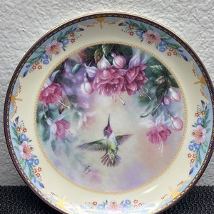 May include: A decorative porcelain plate with a hummingbird in flight against a background of pink and purple flowers. The plate has a gold rim and a blue and white border with floral designs. The artist's signature "LGNA" is visible in the lower right corner.