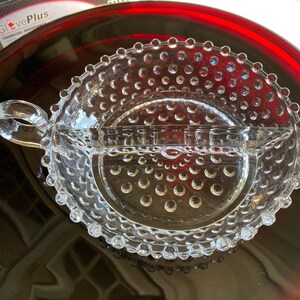 Puede incluir: Un plato dividido de vidrio transparente con asa. El plato tiene un borde con cuentas y una superficie texturizada con pequeños puntos en relieve.