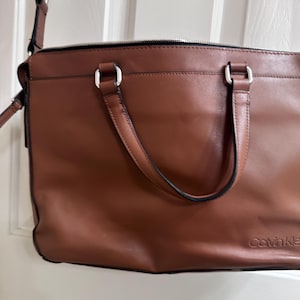 May include: A brown leather Calvin Klein shoulder bag with a silver zipper and handles. The bag has a black shoulder strap and the Calvin Klein logo is visible on the lower right corner.