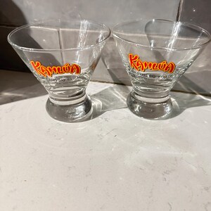 May include: Two clear Kahlúa cocktail glasses with a flared bowl and a short stem. The Kahlúa logo is printed in red and yellow on the front of each glass. The text "everyday exotic" is printed below the logo.