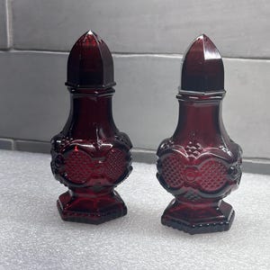 Puede incluir: Dos saleros y pimenteros a juego de cristal rojo rubí. Cada salero y pimentero tiene un diseño decorativo y texturizado alrededor del cuerpo y una parte superior puntiaguda. Los saleros y pimenteros se muestran sobre una superficie blanca.