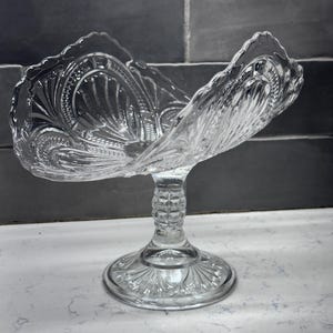 May include: A clear, pressed glass pedestal bowl with a scalloped rim. The bowl has an ornate, textured design and sits on a round base. The stem has a textured, vertical pattern.