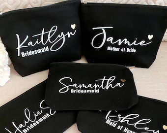 May include: Five black zippered pouches with white lettering and gold hearts. The pouches are personalised with the names Kaitlyn, Jamie, Samantha, Hailie, and Ashley. The pouches are labelled with the titles "Bridesmaid", "Mother of Bride", "Maid of Honor", and "Bridesmaid".