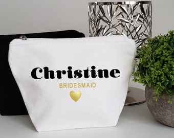 May include: White canvas makeup bag with black text that reads "Christine Bridesmaid" and a gold heart. The bag is sitting on a white surface next to a black makeup bag.