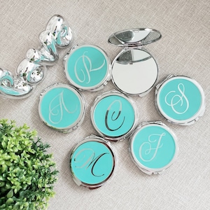 May include: Set of seven silver compact mirrors with turquoise blue lids. Each mirror has a different letter of the alphabet engraved on the lid in a silver script font.