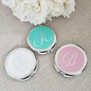 May include: Three compact mirrors with silver frames and different colored lids. The lids are turquoise, white, and pink. Each lid has a silver letter, A, B, and B, in a cursive font.