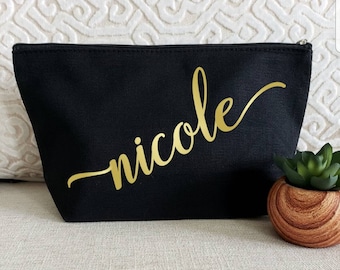 May include: A black canvas cosmetic bag with a gold script font that reads "nicole". The bag has a zipper closure and is sitting on a neutral-colored surface. A small succulent in a wooden pot is next to the bag.