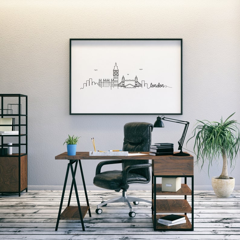 May include: A black framed picture of a line drawing of the London skyline, including Big Ben and Tower Bridge, with the word "london" written in cursive. The picture is hanging on a white wall above a wooden desk with a black office chair, a potted plant, and a desk lamp.