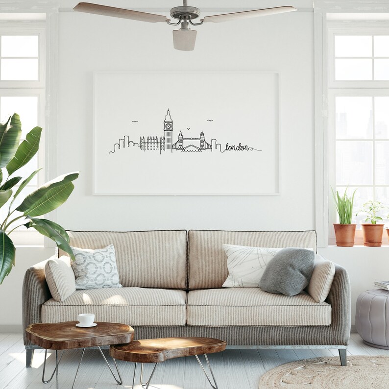 May include: A black and white line drawing of the London skyline, including Big Ben, the London Eye, and Tower Bridge. The drawing is framed and hanging on a white wall above a beige sofa with pillows. The word "london" is written in cursive below the skyline.