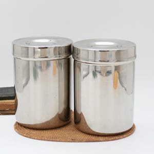 Peut inclure: Deux boîtes de conservation cylindriques en acier inoxydable avec des surfaces polies et réfléchissantes et des couvercles plats assortis. Les récipients présentent un design lisse et sans couture, avec une finition argentée métallique.