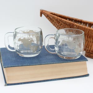 Puede incluir: Dos tazas de vidrio transparente con asas, cada una con un diseño de mapa del mundo, están sobre un libro azul. Una cesta de mimbre está en el fondo. Las tazas son ideales para café o té.
