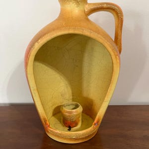 May include: A ceramic wall-mounted candle holder in the shape of a jug. The holder is primarily yellow with orange and brown accents. It features a handle and a small, round candle cup at the base. The jug has a crackled glaze.
