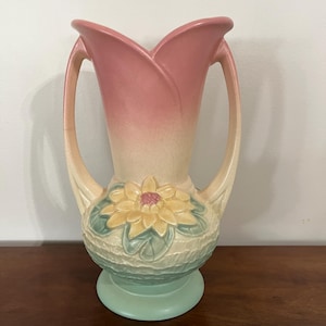 Puede incluir: Un jarrón de cerámica vintage con una parte superior en forma de tulipán, asas y un diseño floral. El jarrón presenta un degradado de colores rosa y crema, con una base verde y un diseño floral amarillo y rosa.