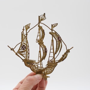 May include: A detailed, golden filigree ship sculpture with three masts and delicate rigging. The ship features intricate openwork designs, small flags, and a cross emblem on the sails. A decorative nautical piece.