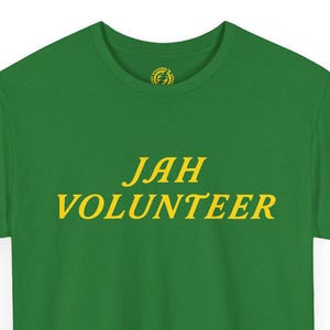 May include: A green short-sleeved t-shirt featuring the text JAH VOLUNTEER printed in yellow across the chest. The shirt has a crew neckline and a small yellow circular logo graphic located on the inside of the collar area at the back neck.