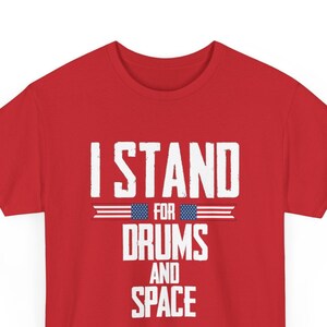 May include: Red t-shirt with white text that reads "I Stand For Drums And Space". The text is framed by two horizontal lines with an American flag design.
