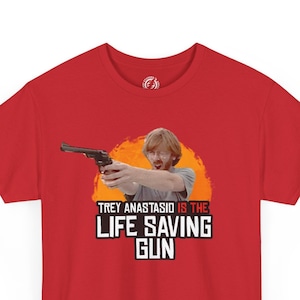 May include: A red t-shirt featuring a graphic of a man holding a gun, with the text "TREY ANASTASIO IS THE LIFE SAVING GUN" in white and orange. The shirt has short sleeves and a crew neck.