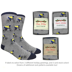 May include: Gray socks with a navy blue cuff and toe, featuring a repeating pattern of cyclists in yellow and blue. Three gift boxes display socks with holiday greetings and names.
