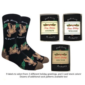 May include: A pair of black socks with a sloth design and the text "Don't Hurry Be Happy". The socks are packaged in a box with a personalized holiday greeting label. The label has a green wreath with red berries and the text "Merry Christmas ANDREW".