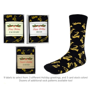 May include: Three gift boxes with personalized holiday greetings and names, "Zachary", "Ryan", and "David". Each box contains a pair of black socks with a gold trumpet pattern. The socks are displayed on a white background.