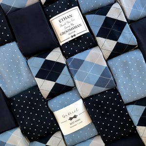 May include: An assortment of rolled socks in various shades of blue and navy, featuring argyle, polka dot, and solid patterns. Some socks have labels that read "Ethan Thank You For Being My Groomsman 04.11.26" and "Gabriel Best Man November 7, 2026".